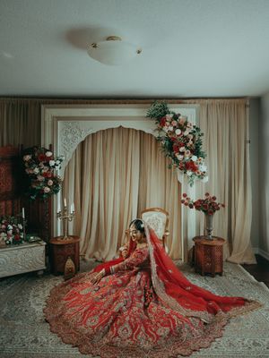 Indian bride in ornate red lehenga seated before floral arch backdrop in elegant bridal portrait.