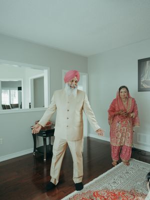 Sikh man in pink turban and cream sherwani with woman in red salwar suit in home setting.