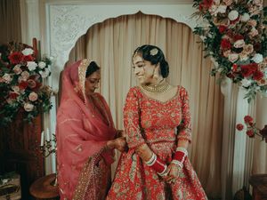 Indian bride in red lehenga shares tender moment with elder woman in pink dupatta at wedding.