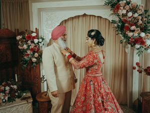 A Sikh bride in a red embroidered lehenga adjusts the sherwani of an elderly man in a pink turban.