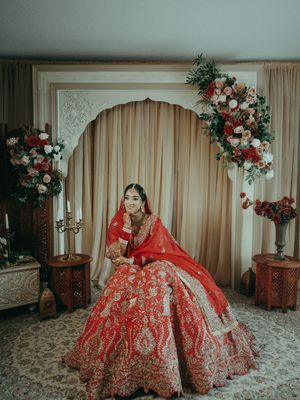 Indian bride in ornate red lehenga posing under decorative arch with floral arrangements at wedding.