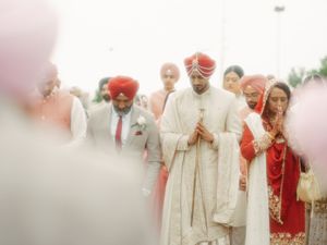 Sikh groom in white sherwani with red turban walks in ceremonial procession surrounded by family in traditional attire.