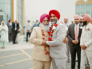 Two Sikh men in turbans embrace warmly outdoors at wedding ceremony with guests in background.