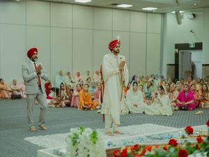Sikh groom in white sherwani and red turban stands during Anand Karaj wedding ceremony in a gurdwara hall.