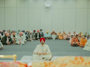 Sikh groom in red turban and white sherwani seated during Anand Karaj ceremony with guests in background.