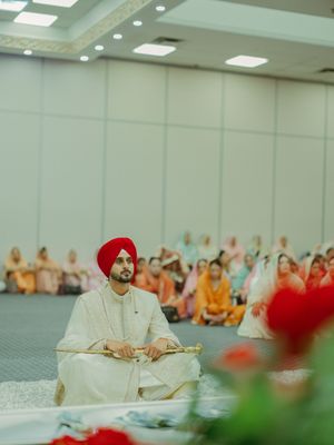 Sikh groom in white sherwani and red turban sitting during Anand Karaj wedding ceremony in decorated hall.