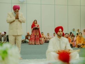 Sikh wedding ceremony with bride in red lehenga, groom in pink turban, and guests seated in a decorated hall.