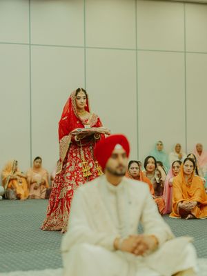 Sikh bride in red lehenga carries ceremonial tray during Anand Karaj wedding ceremony while groom sits in white sherwani.