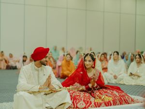 Sikh bride and groom in traditional red and white attire during Anand Karaj wedding ceremony indoors.