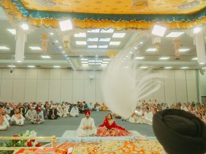 Sikh wedding ceremony in a decorated hall with bride and groom seated before Guru Granth Sahib, chaur sahib being waved above.