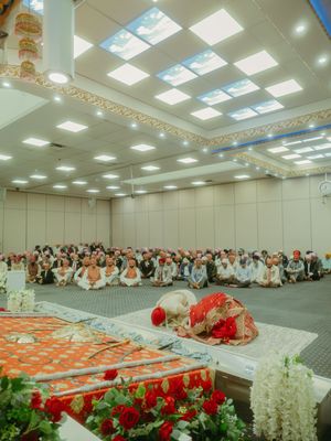 Sikh wedding ceremony in a gurdwara hall with bride and groom bowing before the Guru Granth Sahib surrounded by seated guests.