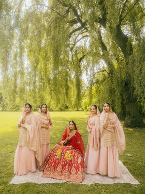 Bride in red lehenga surrounded by bridesmaids in pink outfits under a weeping willow tree.