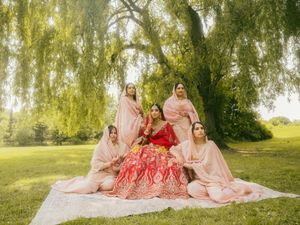 Indian bride in red lehenga poses with bridesmaids in pink under lush weeping willow tree.