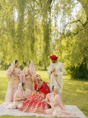 Sikh bride and groom with bridesmaids in pink seated on blanket under weeping willow tree.