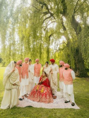 Sikh groom in pink turban surrounds bride in red lehenga with groomsmen under willow tree.