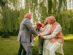 Sikh men in red turbans and traditional attire greet each other joyfully outdoors under a weeping willow tree.
