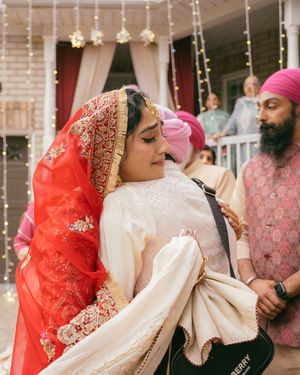 Sikh bride in red dupatta and white outfit embraces a guest during outdoor wedding celebration with decorated entrance.