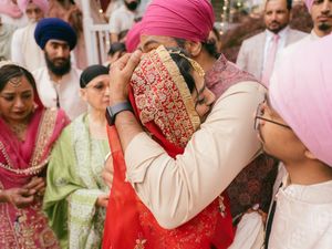 Sikh wedding ceremony embrace between bride in red and groom in pink turban, surrounded by guests in traditional attire.