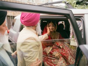Sikh groom in pink turban touches brides face as she sits in decorated car in traditional red bridal outfit.