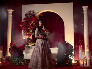 Woman in embroidered gray lehenga poses before red floral arch with roses and candles at Indian wedding.