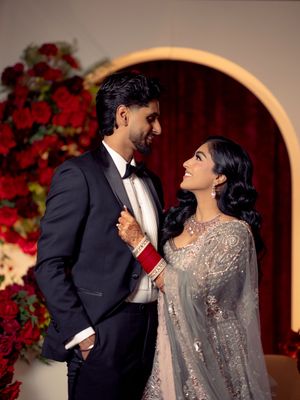 A couple in elegant wedding attire gazes lovingly at each other before a red floral arch backdrop.
