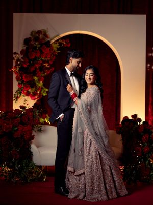 Couple in elegant wedding attire pose before floral arch with red roses at Indian reception.