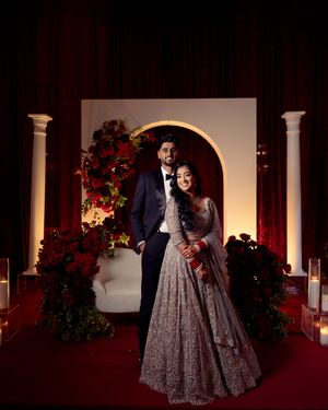Couple in elegant Indian wedding attire pose before a white arch with red floral decor and candles.