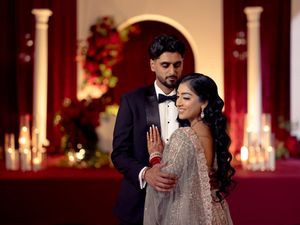 South Asian couple in elegant wedding attire pose romantically against a candlelit red backdrop.