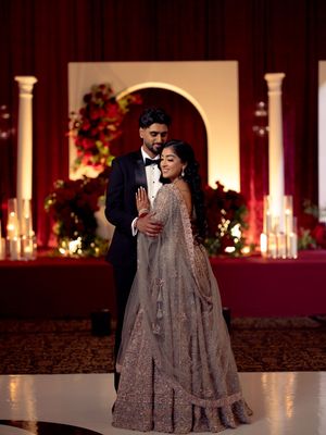 Bride in gray lehenga and groom in tuxedo share romantic dance at candlelit Indian wedding.