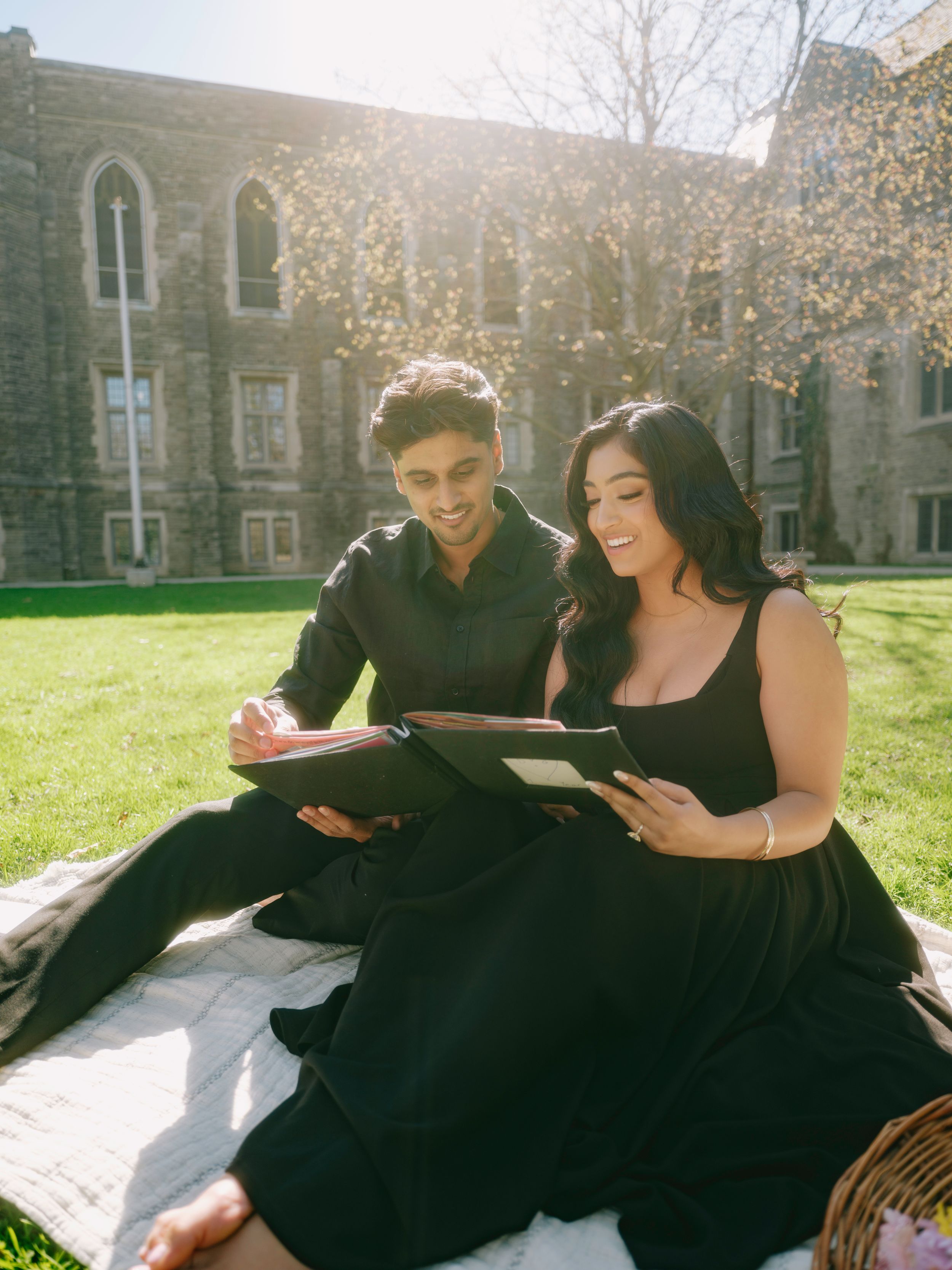 Couple in black outfits sitting on blanket outdoors, looking at tablet together near stone building on sunny day.