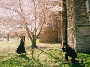 Couple posing outdoors near a stone building with blooming pink cherry blossom tree on a sunny spring day.