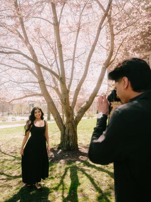 Man photographing woman in black dress under blooming cherry blossom tree during spring engagement session.