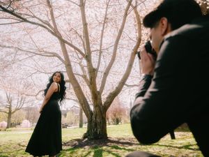 Man photographing woman in black dress standing under blooming cherry blossom tree in spring park.