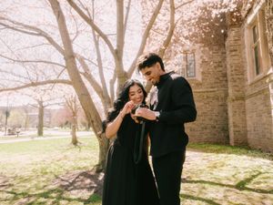 Couple in black formal wear sharing a tender moment under cherry blossom trees near stone building.