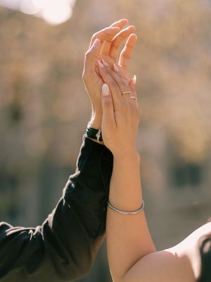 Two hands reaching up together, one wearing a ring and bracelet, against a blurred background.
