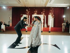 Couple sharing first dance at Indian wedding reception with sparklers and red drape backdrop on white dance floor.