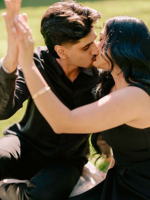 Couple dressed in black sharing a romantic kiss outdoors in sunlit garden during engagement photo session.