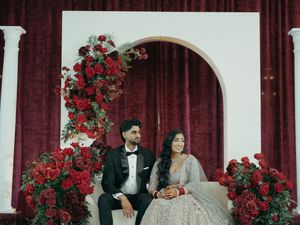 Bride and groom seated before a white arch adorned with red roses at an elegant South Asian wedding reception.
