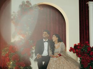 South Asian couple in wedding attire smiling joyfully, groom in tuxedo, bride in gold lehenga, surrounded by red roses.