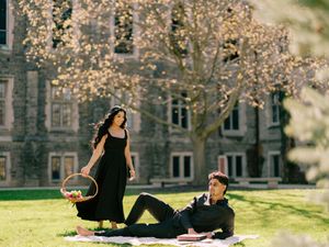 Couple enjoying a spring picnic on green lawn before a stone building, woman in black dress holding basket.