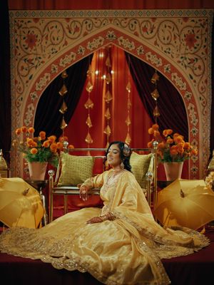A bride in golden bridal attire sits regally before an ornate Mughal-style arch with marigold flowers and rich red drapes.
