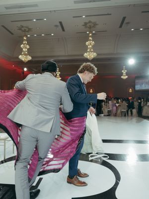 Two men in suits handle a large pink fabric on a dance floor at an elegant wedding reception venue.
