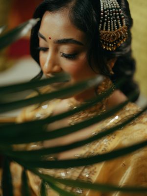 A South Indian bride in a golden saree with ornate hair jewelry, framed by tropical leaves.