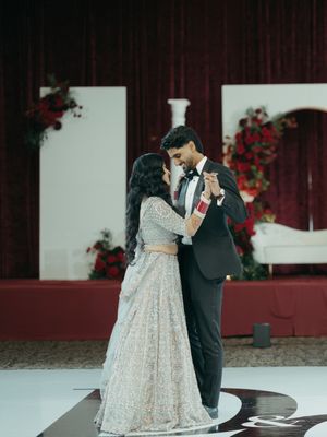 Couple shares first dance in elegant wedding attire, bride in embellished lehenga, groom in black tuxedo, red floral backdrop.