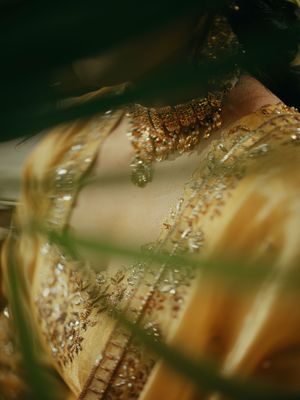 Close-up of a bride in golden embroidered lehenga with ornate gold necklace, shot through green foliage.