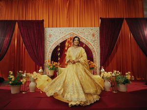 A bride in a golden yellow lehenga twirls before an ornate arch backdrop with red drapes and floral arrangements.
