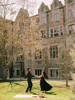 Couple in black outfits dancing on grass in front of a historic stone building during a sunny spring day.