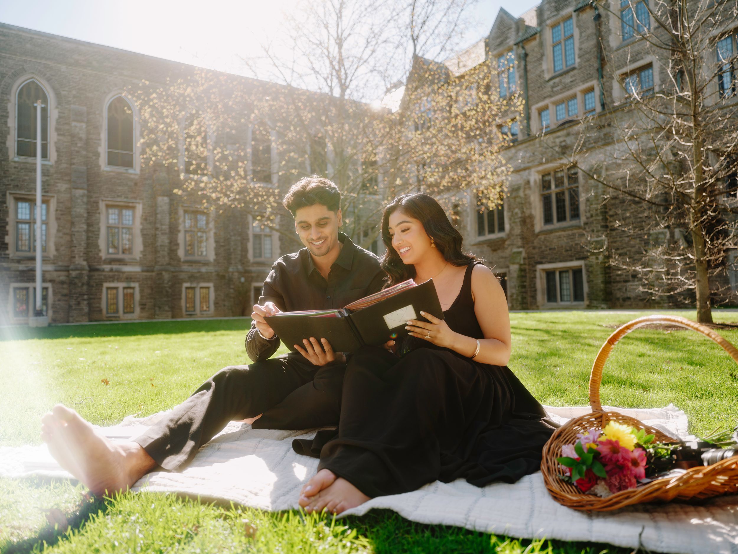 Two students in black outfits share a tablet during a sunny campus picnic on green grass near a stone university building.