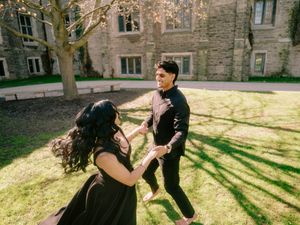 Young couple holding hands and spinning joyfully on green lawn beside a gothic stone university building.