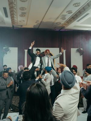 Two men lifted by celebrating guests at a Sikh wedding reception in an elegant banquet hall.